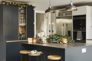 dark gray kitchen with festive decorations on kitchen island, fir tree, candles, walnuts, cloches with baubles in, bar stools, drinks cabinet