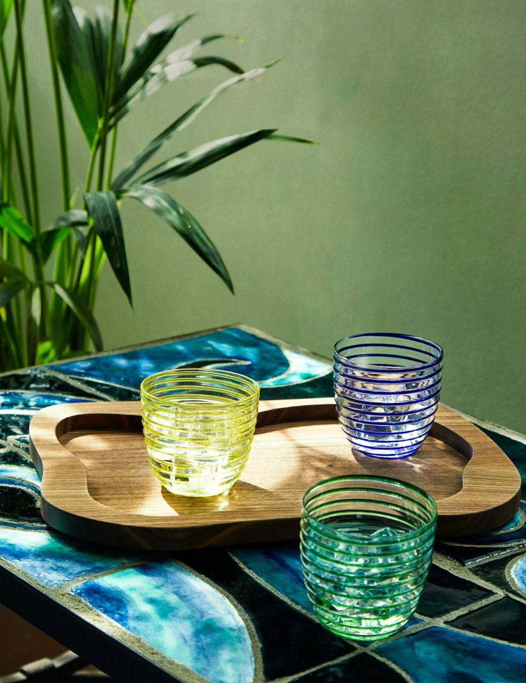 A Moribana Walnut Tray holding two colorful glasses in yellow and blue glass, both of which sit atop a patterned blue ceramic and wrought iron table together with the tray and another green glass in a naturally lit space with plants.