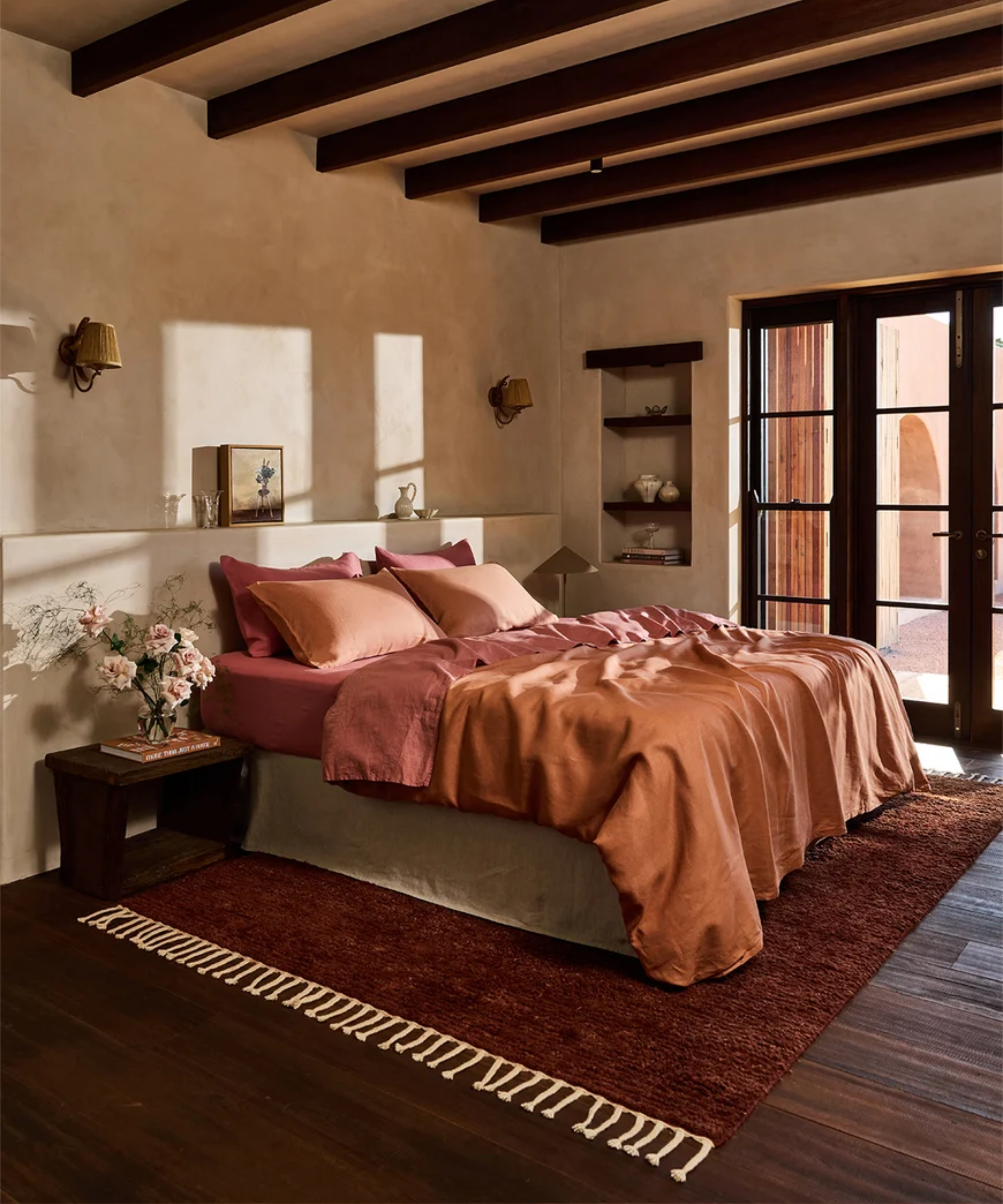 Orange-and-rose layered bed sheets styled atop of a skirted bed in a Spanish-style bedroom with large windowpane doors and rustic wood beams overhead.