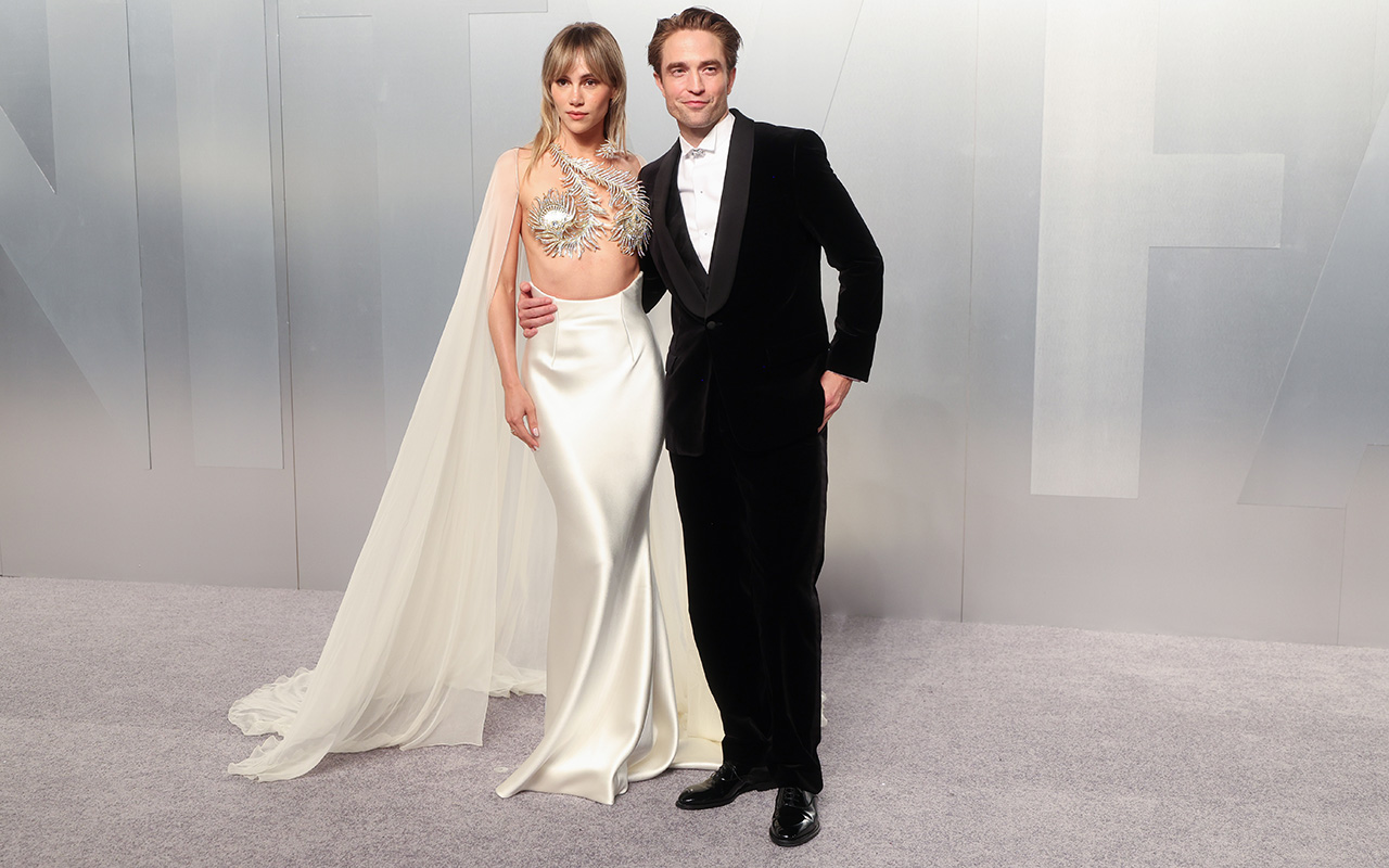 Full look at Suki Waterhouse's satin gown with feather top and gauzy cape. She's standing next to Robert Pattinson in a classic tux.