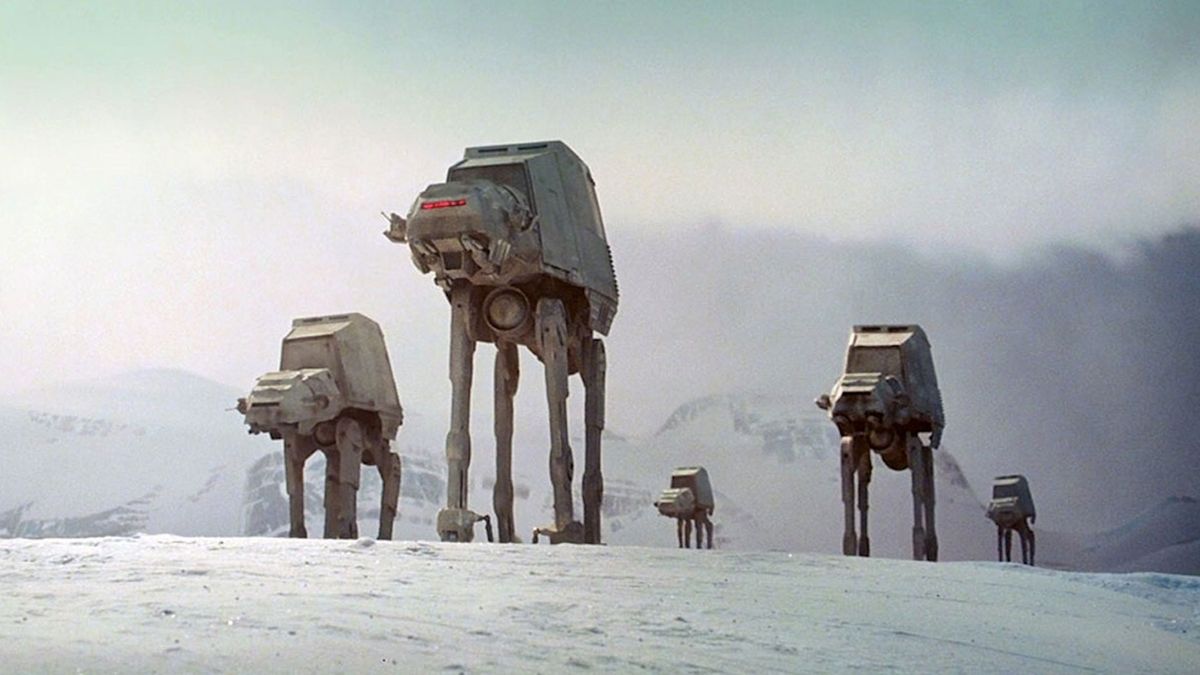A series of Grey camel-like robots are walking on snow with mountains in the background.
