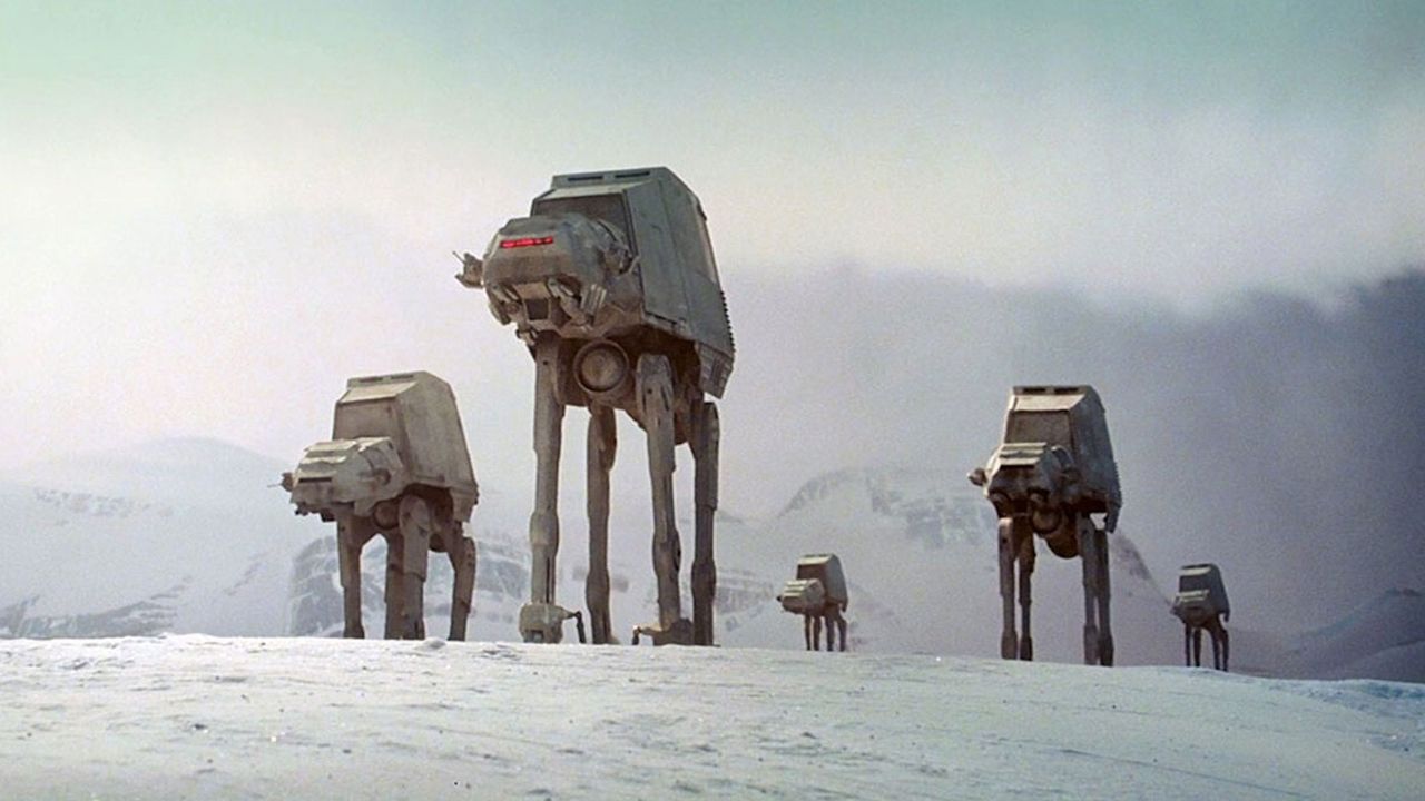 A series of Grey camel-like robots are walking on snow with mountains in the background.