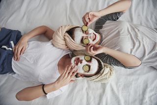 Two women lying on a bed wearing avocado face masks.