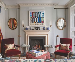 Living space with off white walls and fireplace with two red accent chairs and scalloped ottoman and modern colourful artwork and mirrors