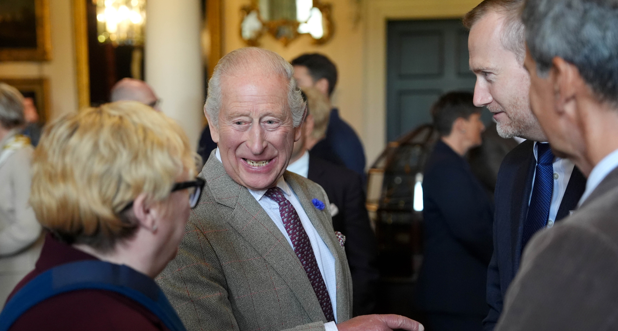 King Charles wearing a gray suit and laughing, talking to a woman