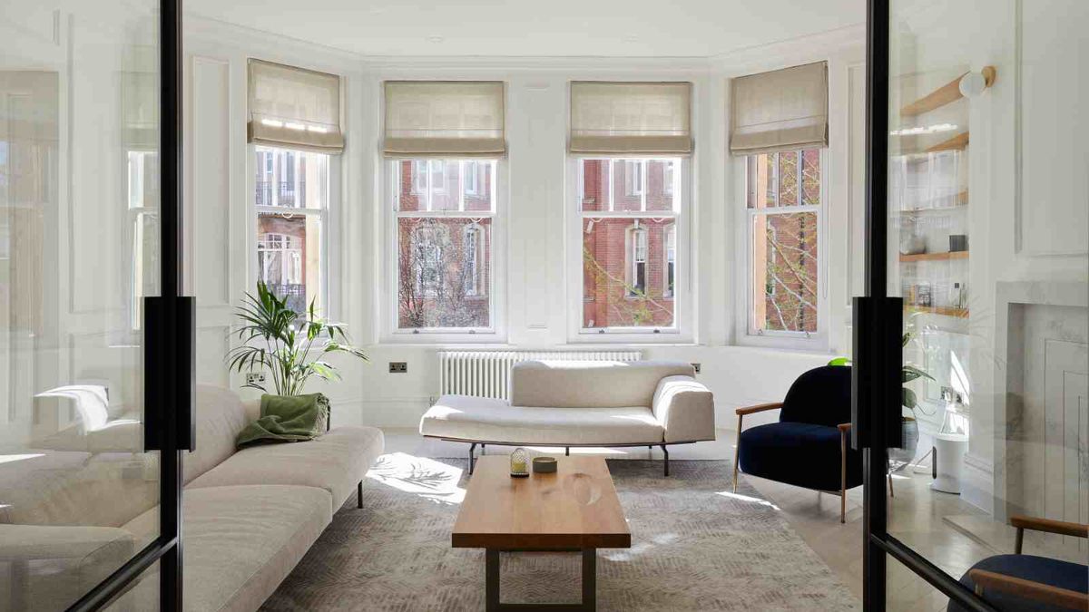 A modern white living room in a period-style home, with four sash windows. Low-level mid-century sofas in white are complemented by two navy armchairs. Two glass doors lead to the room.