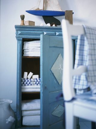 Blue bathroom closet with door open to show towels and soaps in bathroom with armchair and white wall