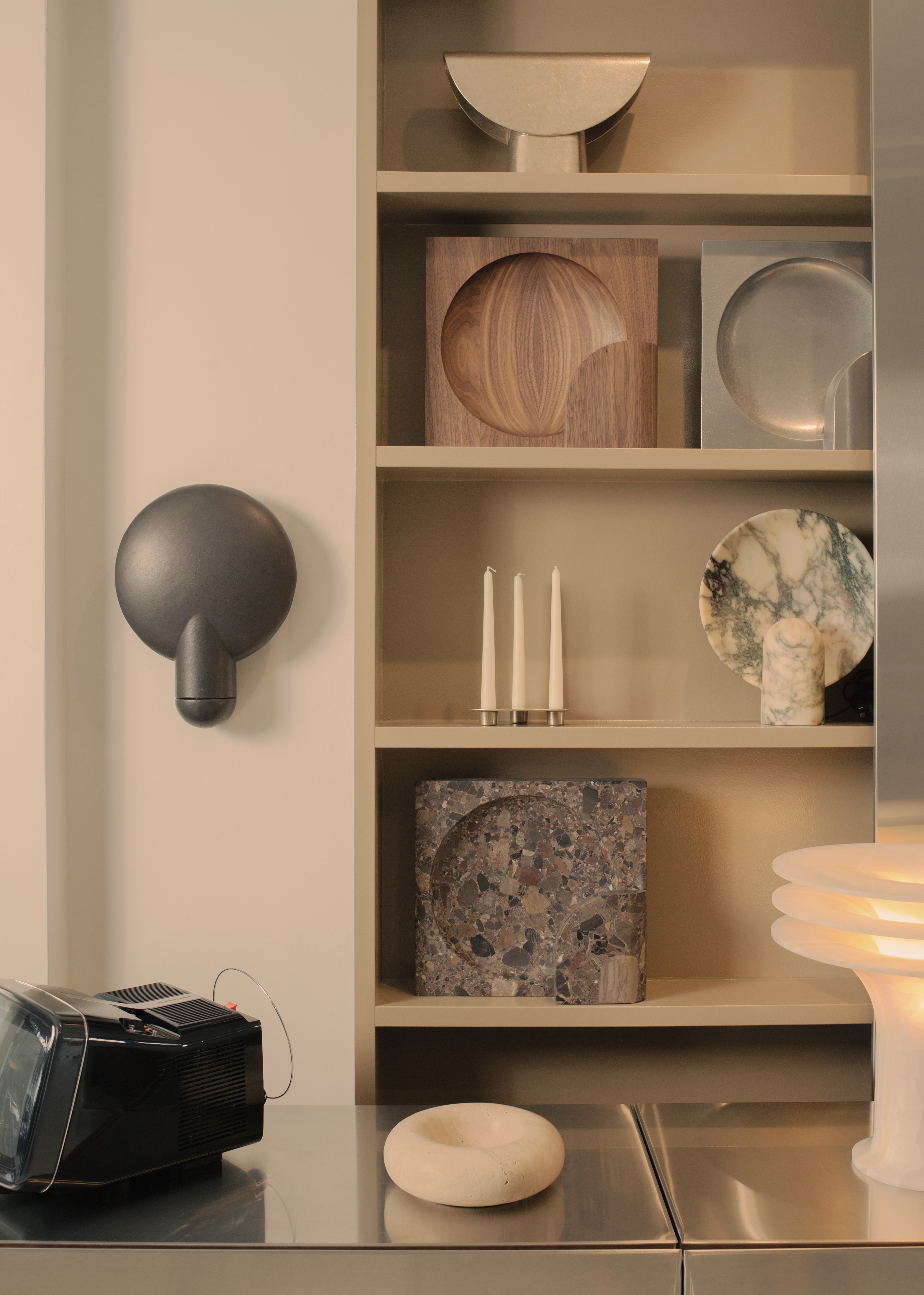 A neutral-tinted showroom features matte chrome surfaces adorned with sculptural collectibles in marble, wood, and silver, and a glowing sculptural lamp.