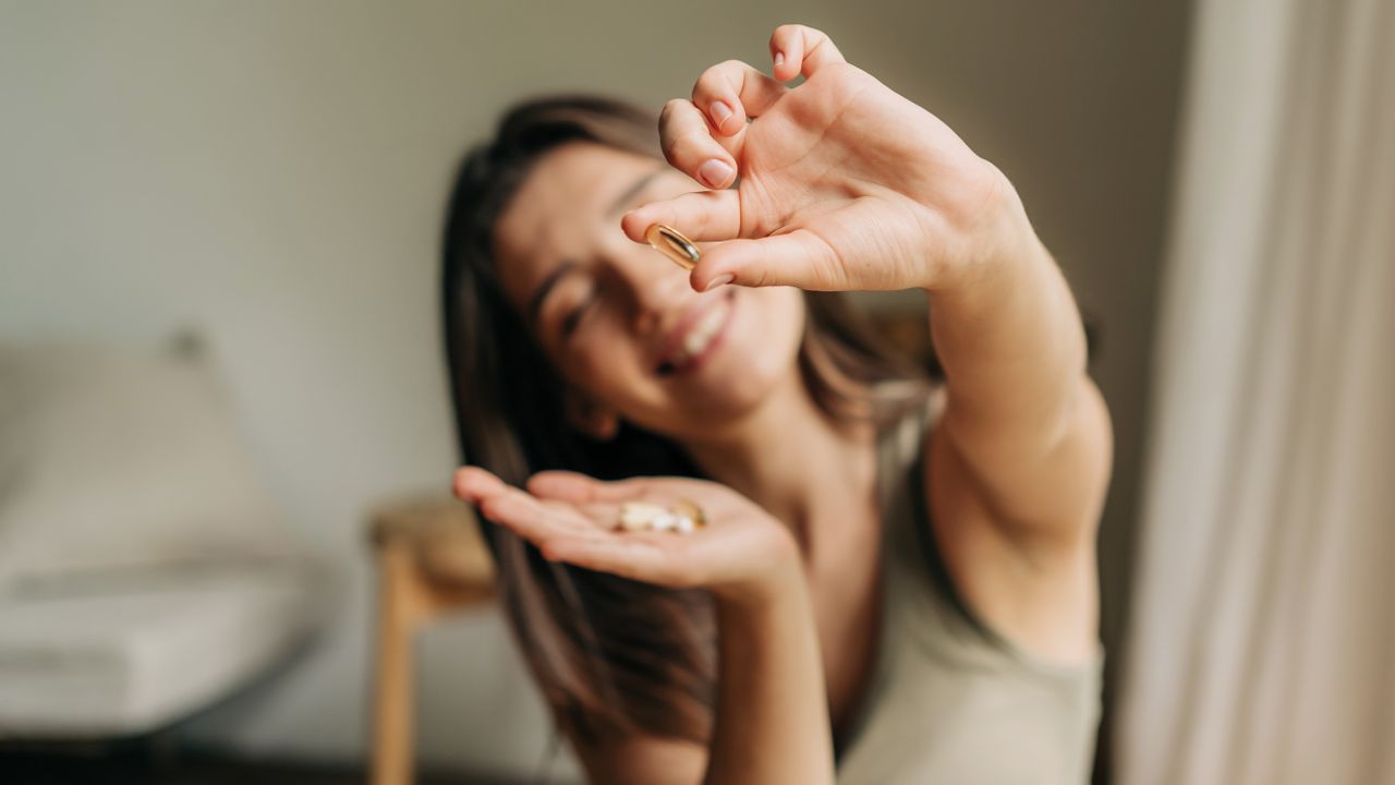 A woman holds a supplement vitamin tablet up towards the camera; she is sitting in a beige living room and wears a vest top