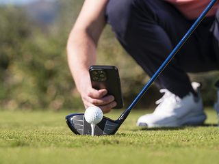 Anders Mankert low down behind the golf ball, taking a picture with his phone of the height of the ball in relation to the clubface