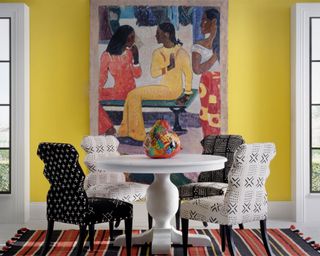 Dining room painted a bright yellow shade, large piece of colorful artwork mounted on wall, sculptural, rounded white dining table, four black and white upholstered dining chairs with cross patterns, striped red and black rug beneath table