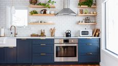 An image of open wooden shelving at eye height with plants, spice racks and cups, and blue kitchen cabinets at knee level in a white subway tiled kitchen 