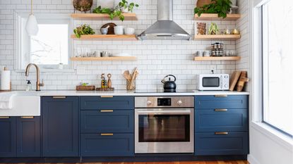 An image of open wooden shelving at eye height with plants, spice racks and cups, and blue kitchen cabinets at knee level in a white subway tiled kitchen 