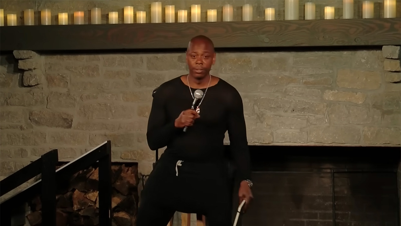 Dave Chappelle Covid special 8:46 sitting on a stool with candles behind him.