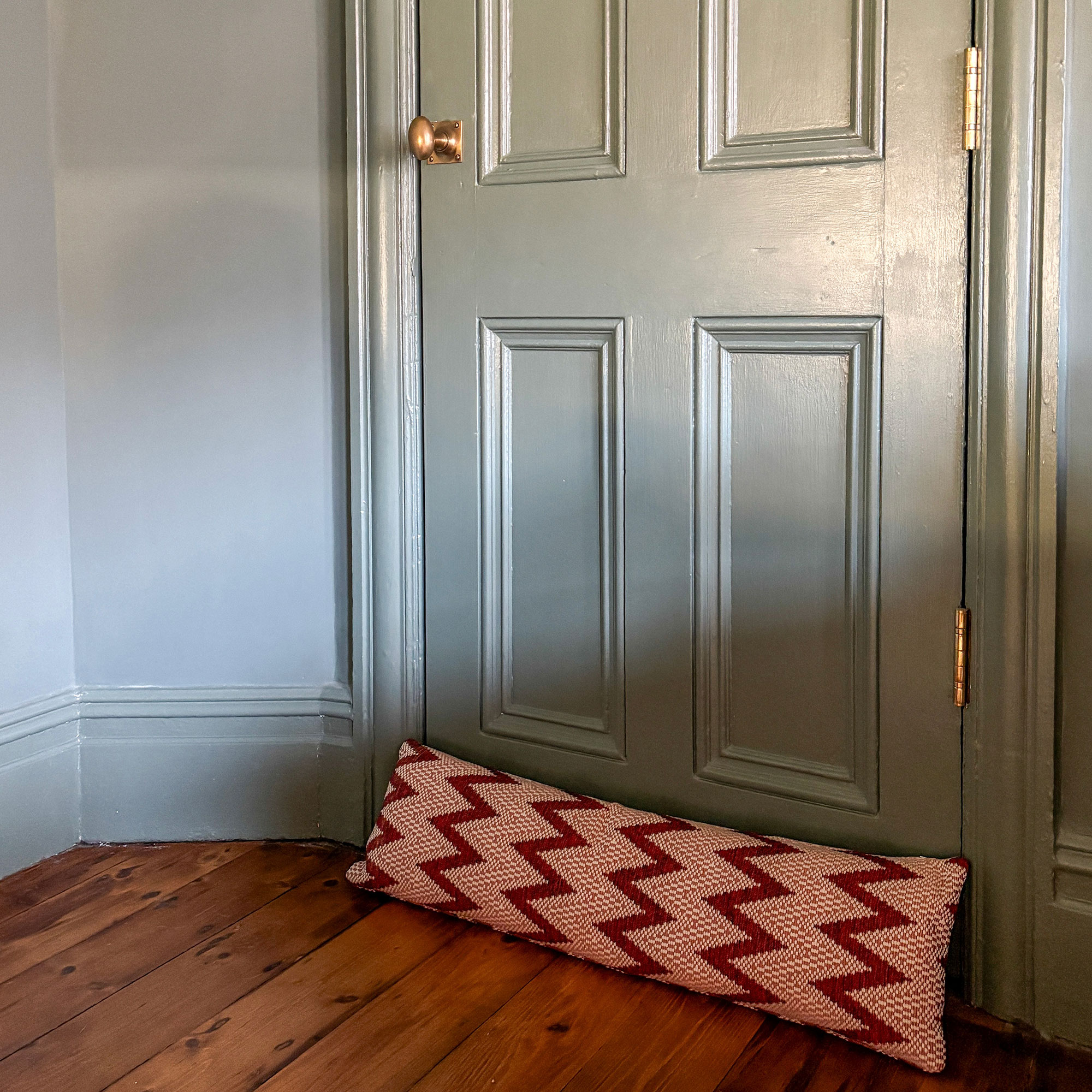 Pink and red zig-zag patterned draught excluder on wooden floor beside green-grey door