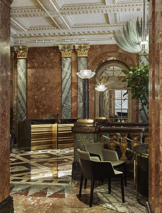 The lobby in the Mandarin Oriental Hyde Park. A high ceiling painted in white, with gold trimming overlooks a majestic staircase in gold, brown, and shades of gray marble. The feather-like, silver chandelier is in the center, while the floors and walls are all in the same shades of marble as the staircase.