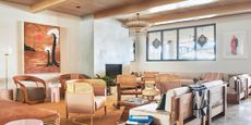 A hotel lounge decorated with earthy, organic furniture, including rattan, wood, and pale pink upholstered armchairs and sofas, terracotta-shaded geometric rugs, and abstract artworks is captured when free from people in plain daylight.
