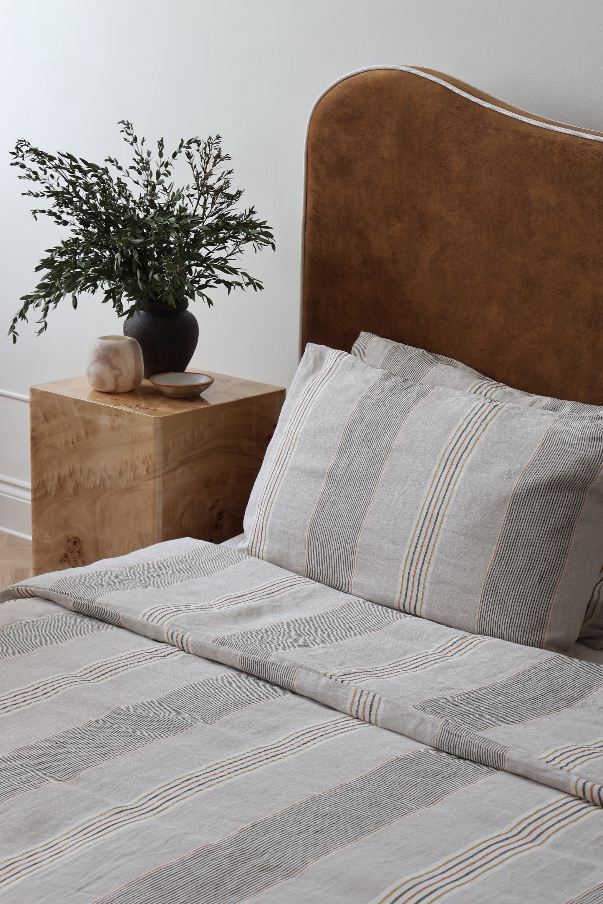striped linen bedding on a tan bed with a burl side table and foliage in a vase
