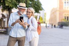 A senior couple standing closely together, focusing on a smartphone in a lively city square, captures a moment of curiosity and exploration amid urban surroundings.
