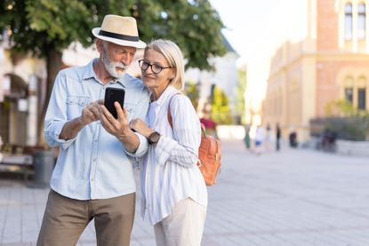 A senior couple standing closely together, focusing on a smartphone in a lively city square, captures a moment of curiosity and exploration amid urban surroundings.
