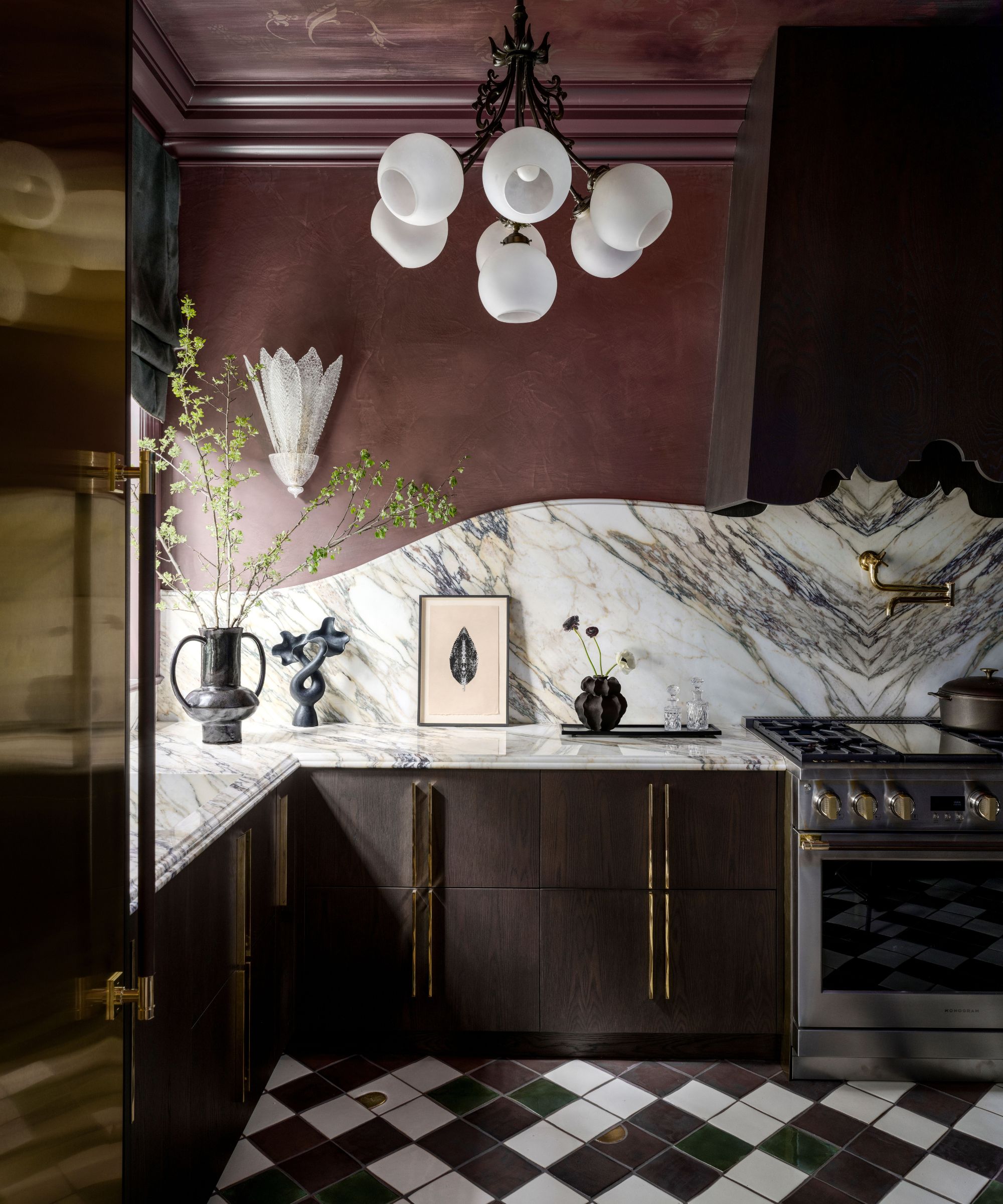 Dramatic kitchen with burgundy walls, heavily veined marble backsplash, dark wood cabinets with brass handles, a globose chandelier, and checkered floor tiles