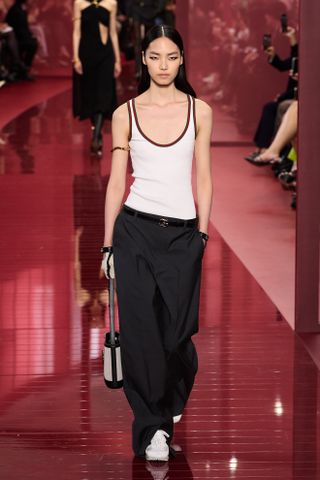 A woman wears a white tank top, black trousers, white sneakers, and a tote bag.