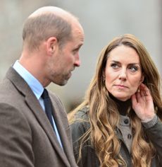 Prince William wearing a brown blazer looking at Princess Kate, putting her hand to her head