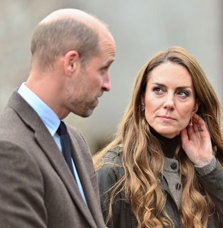 Prince William wearing a brown blazer looking at Princess Kate, putting her hand to her head