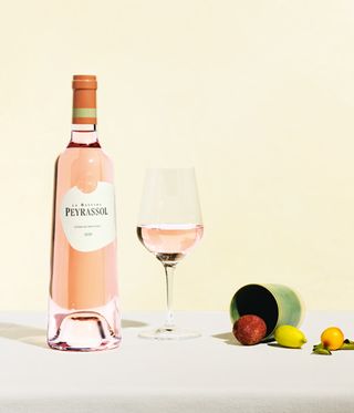A bottle of La Bastide Peyrassol, a partially filled wine glass and fruits against a light pink background