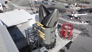 a clam-shell rocket fuselage stands on supports outside a warehouse.
