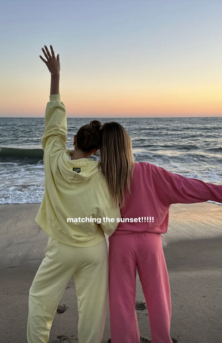 Daily Drills founders Kennedy Crichlow and Mary Ralph Lawson wearing yellow and pink sweat suits on beach.