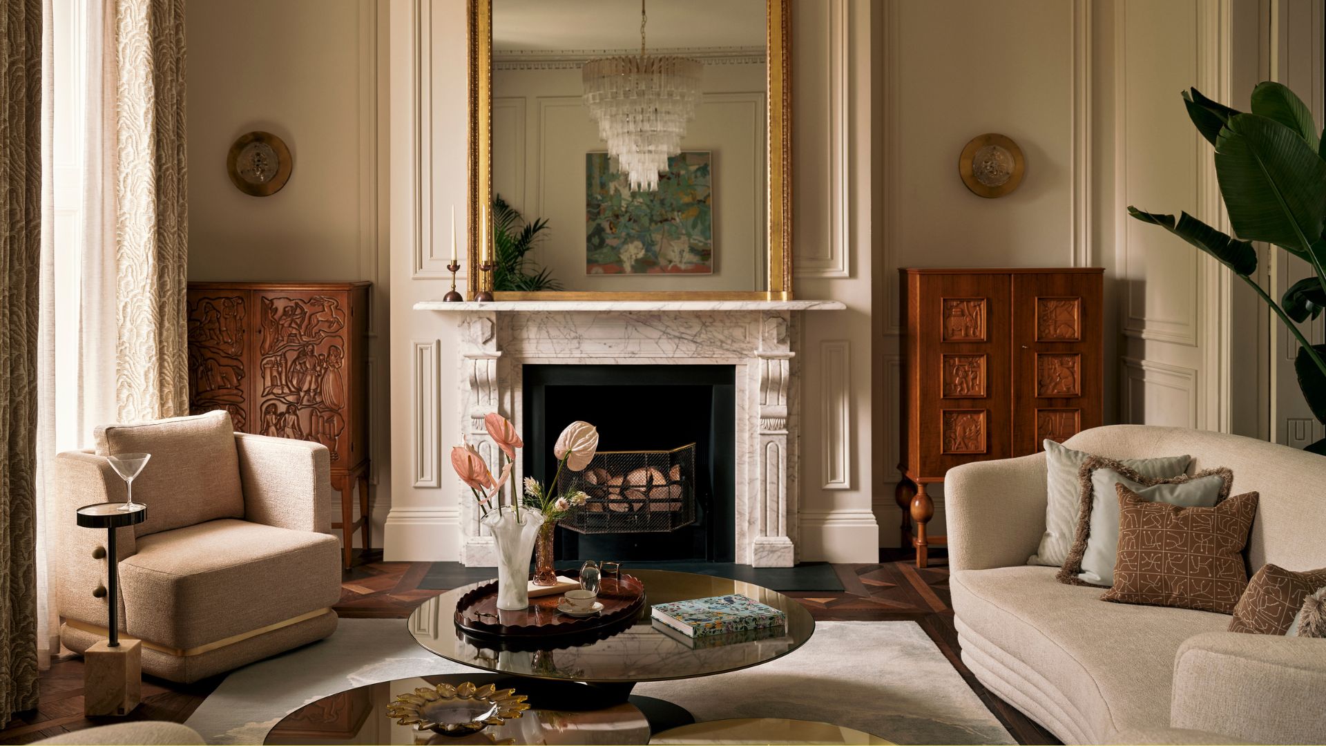 An off-white living room with traditional moulding, a large gold mirror on a marble mantel above a fireplace beside wooden cabinets, a beige sofa, a cream armchair, a side table with a martini glass, and a set of low marble coffee tables with a book and a vase of pink anthuriums