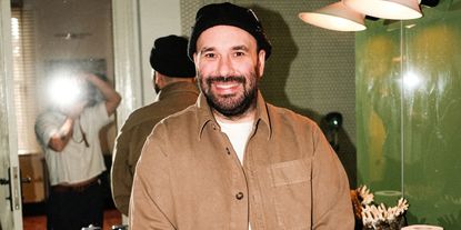 A young man dressed in a white T-shirt, a brown overshirt, and a black beanie smiles at a camera in front of a mirror as his back and the silhouette of a photographer reflect in it.