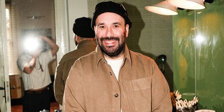 A young man dressed in a white T-shirt, a brown overshirt, and a black beanie smiles at a camera in front of a mirror as his back and the silhouette of a photographer reflect in it.