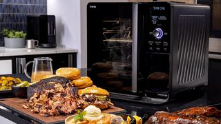 The GE Profile Smart Indoor Smoker on a countertop surrounded by smoked meat, salad, and burger buns