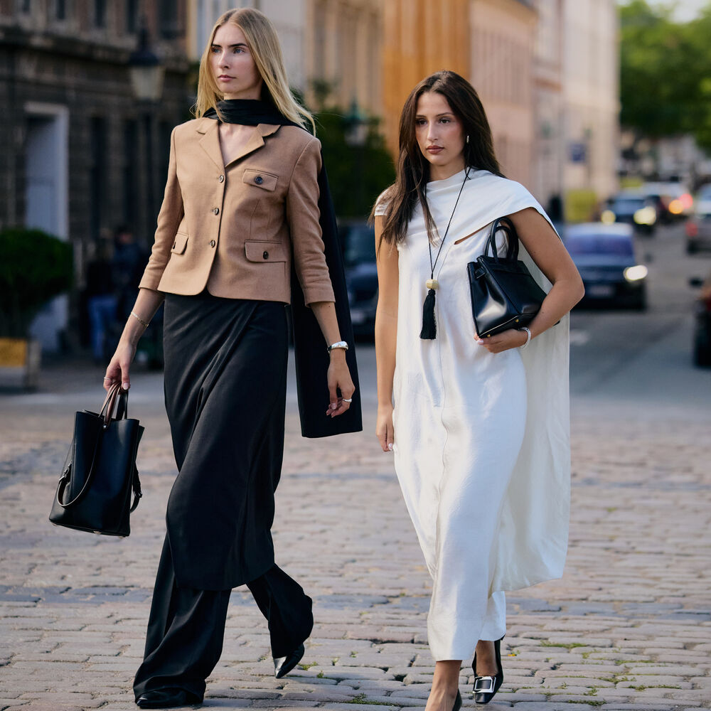 Two women are walking and posing. One wears brown jacket and black pants. The other wears a white dress with heels and black handbag. 