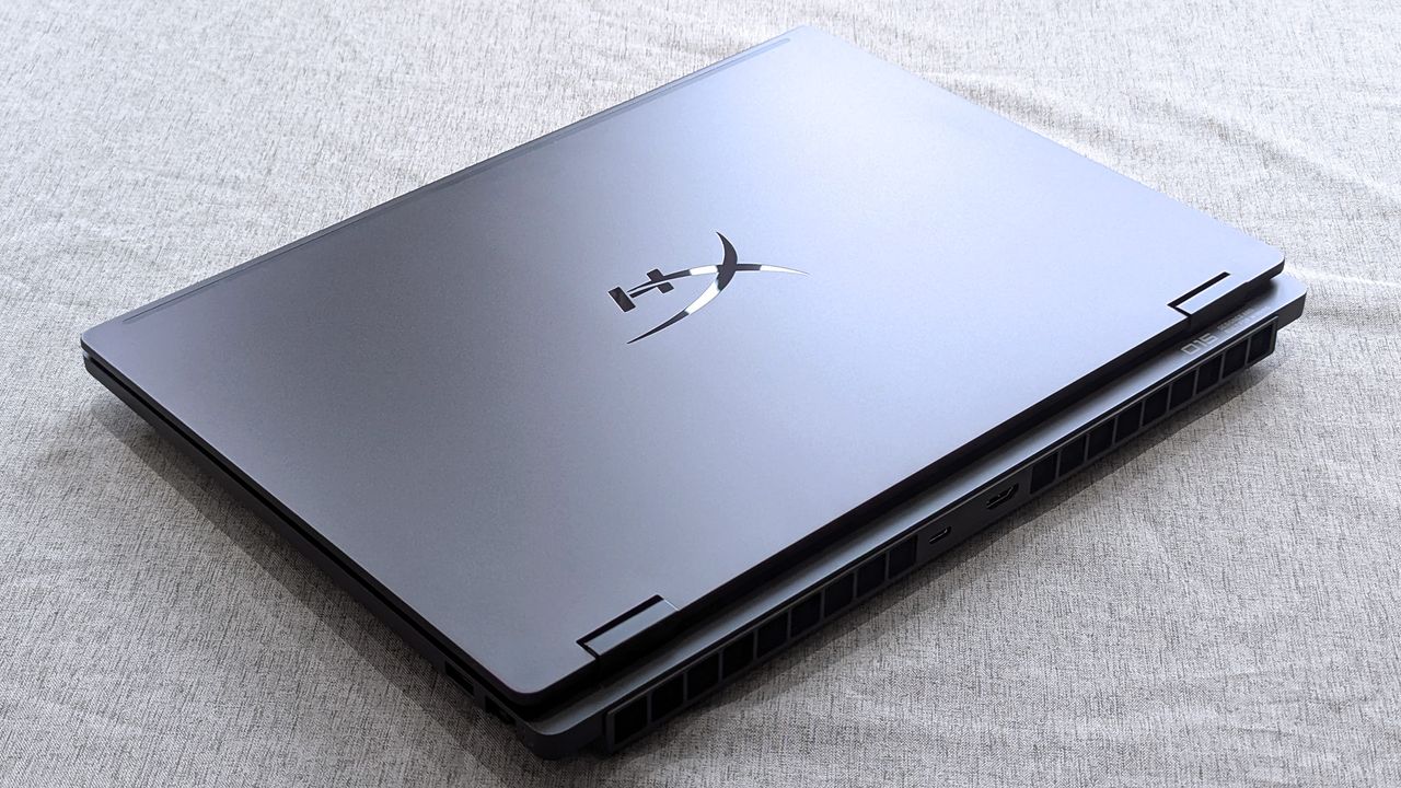 A closed HyperX OMEN 15 laptop resting on fabric, featuring a metallic lid with a reflective HX logo and visible rear ventilation grills along the back edge.
