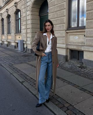 Woman wears white shirt, blue jeans, black boots, brown coat