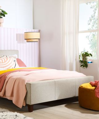 A pink and white bedroom with a light pink wall panel on the left, a gray bed with pink bedding and a yellow throw, a large window with white voile curtains, and a yellow ottoman