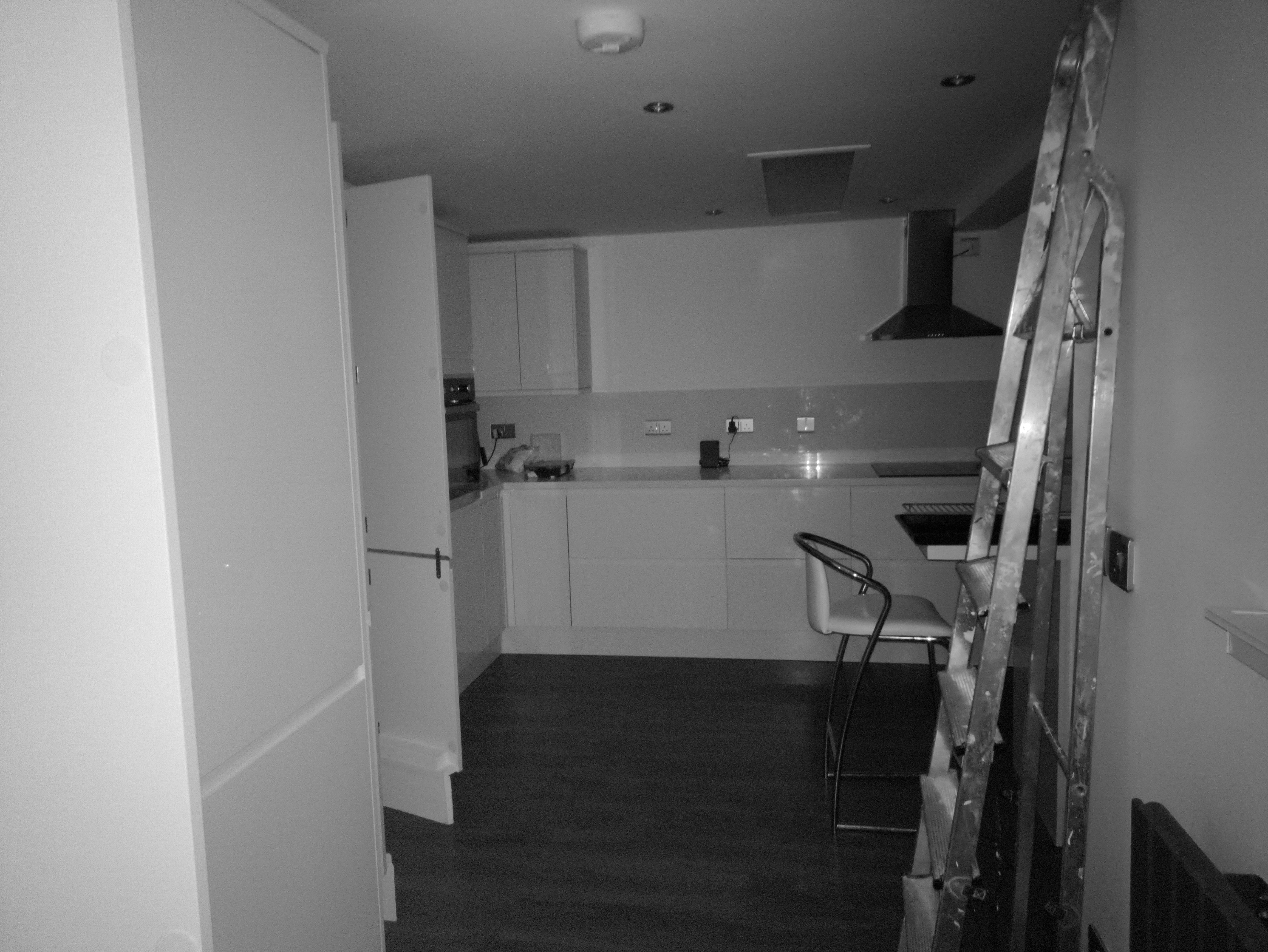 A black and white photo looking down a narrow hallway into a modern kitchen with sleek white cabinets, a black stove hood, and a bar stool next to a tall ladder leaning against the wall.