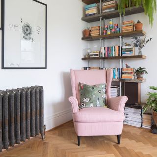 Corner of a living room with a pink armchair in front of a bookshelf filled with books. There's also a metal vintage radiator on a white wall