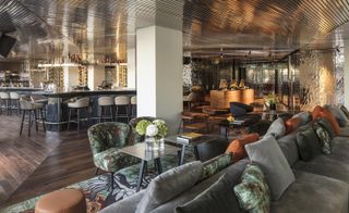 A hotel sitting and dining area with grey sofa's, tables, chairs, a patterned rug, a grey pillar, wooden floors and wooden ceilings.
