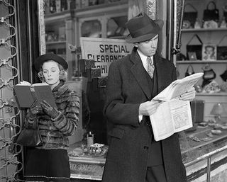 A woman reads a book and a man reads a newspaper outside of a shop in still from the movie The Shop Around the Corner