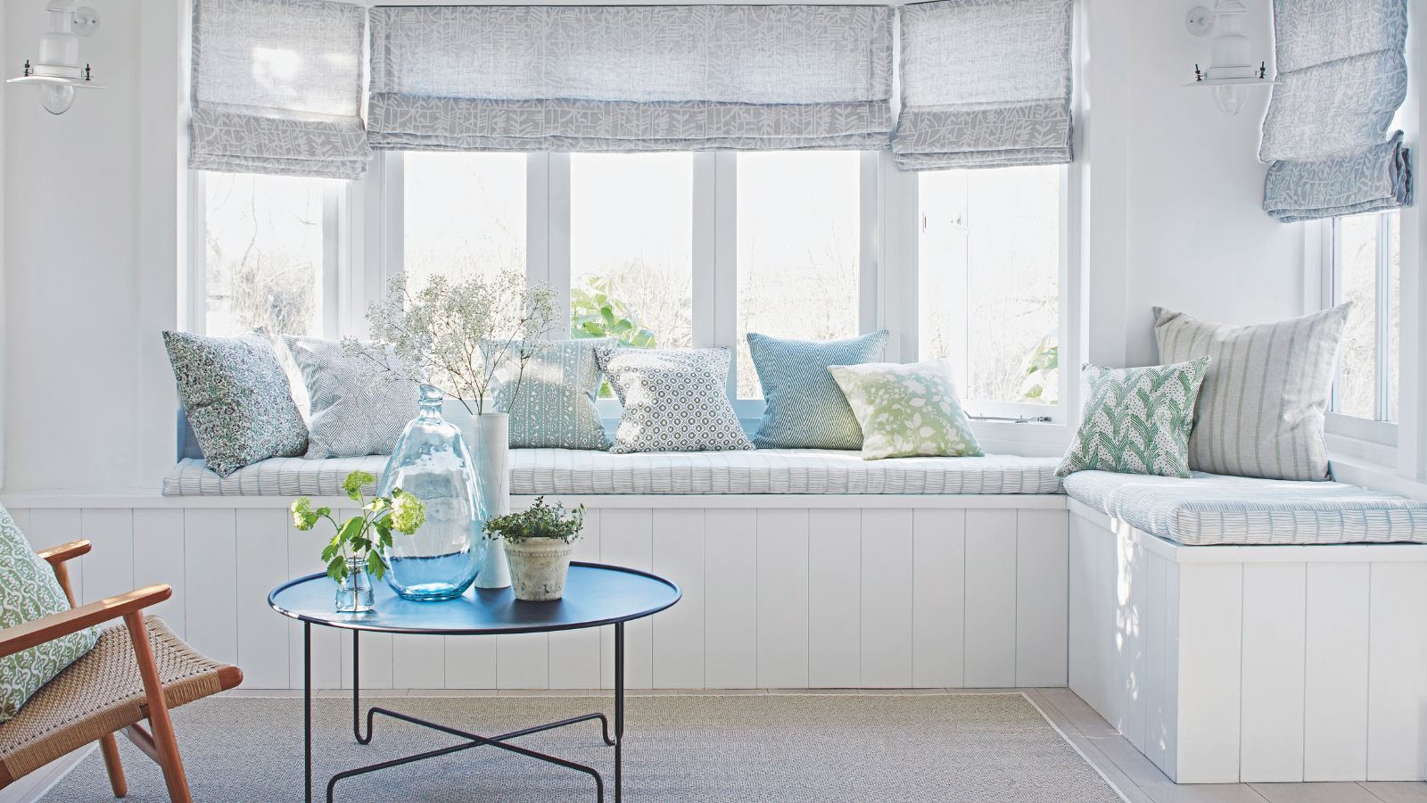 White window nook bench with an assortment of blue and white cushions. It has blue linen window treatments, hessian rug and a coastal chic aesthetic with a mid century chair and metal coffee table in shot