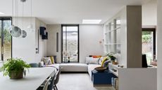 A bright seating area filled with natural light from large windows, with light wooden flooring, a cream corner sofa, and a cream table with glass pendant lights hanging above