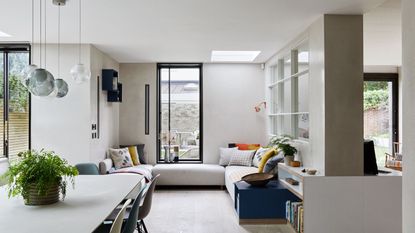A bright seating area filled with natural light from large windows, with light wooden flooring, a cream corner sofa, and a cream table with glass pendant lights hanging above