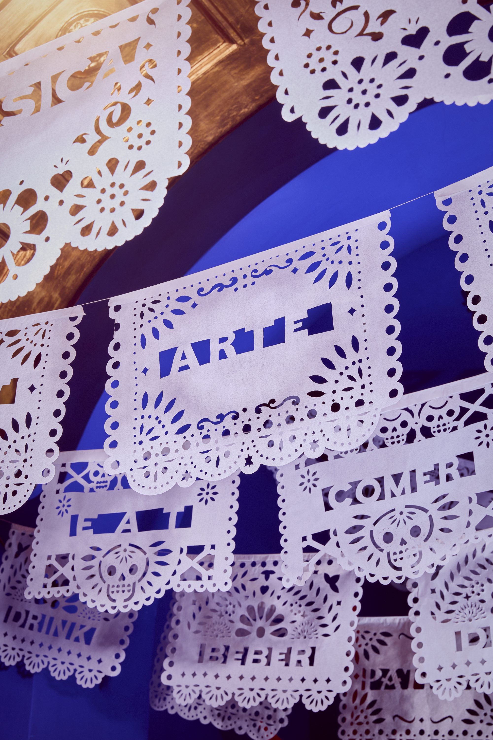 Installation view of a vibrantly colored art show with wicker sculptures resembling cactus, dragons, and sheets of paper bearing different hand-cut messages and hanging from each side of the room.