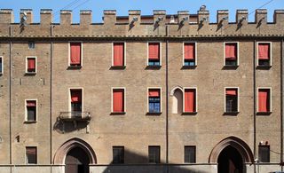 Day time front exterior view of the 14th-century palazzo, brick walled building, windows with red blinds and arched doorways, blue sky