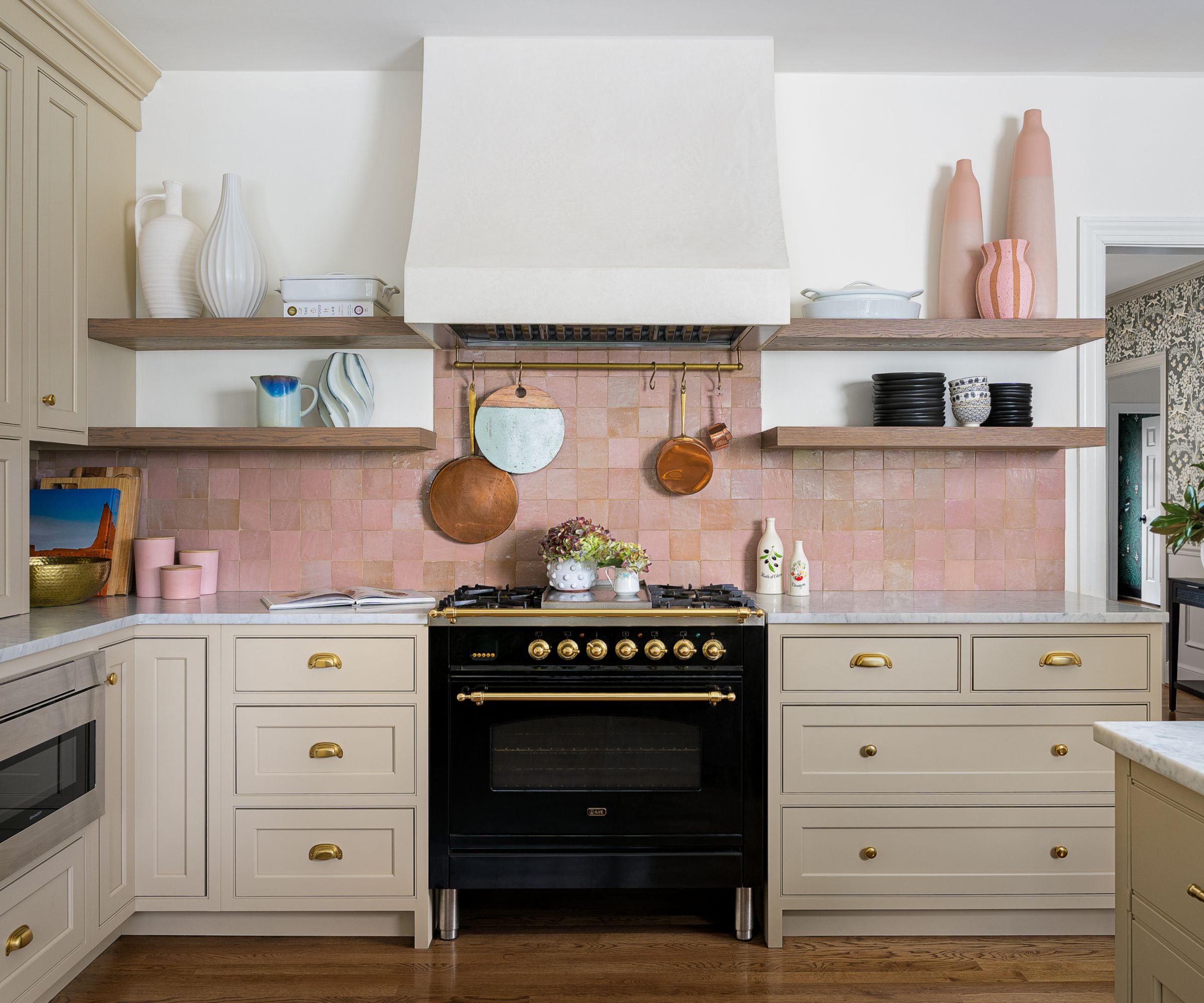 A modern rustic kitchen with beige cabinets, white walls, pink backsplash tiles, brass hardware, and open shelving with decorative objects