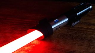 The Darth Vader Force FX Elite Lightsaber sits ignited on a wooden table, with the blade glowing red